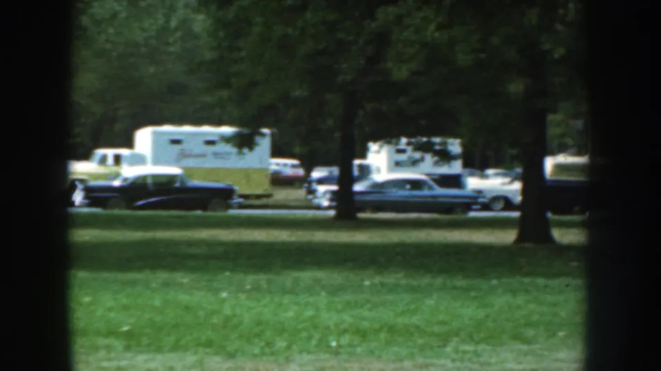 A dark sedan, light-colored cars, and a white and yellow caravan trailer sit along a shaded Nebraska street in 1959 footage.
