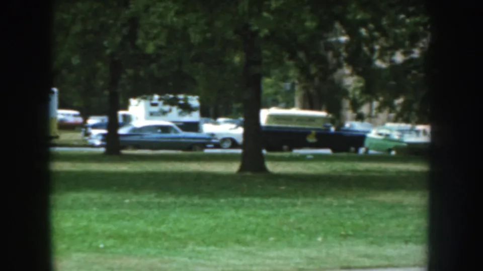 Final view of the suburban street shows a station wagon, dark utility vehicle, caravan trailer, trees, and parked cars in 1959 Nebraska.