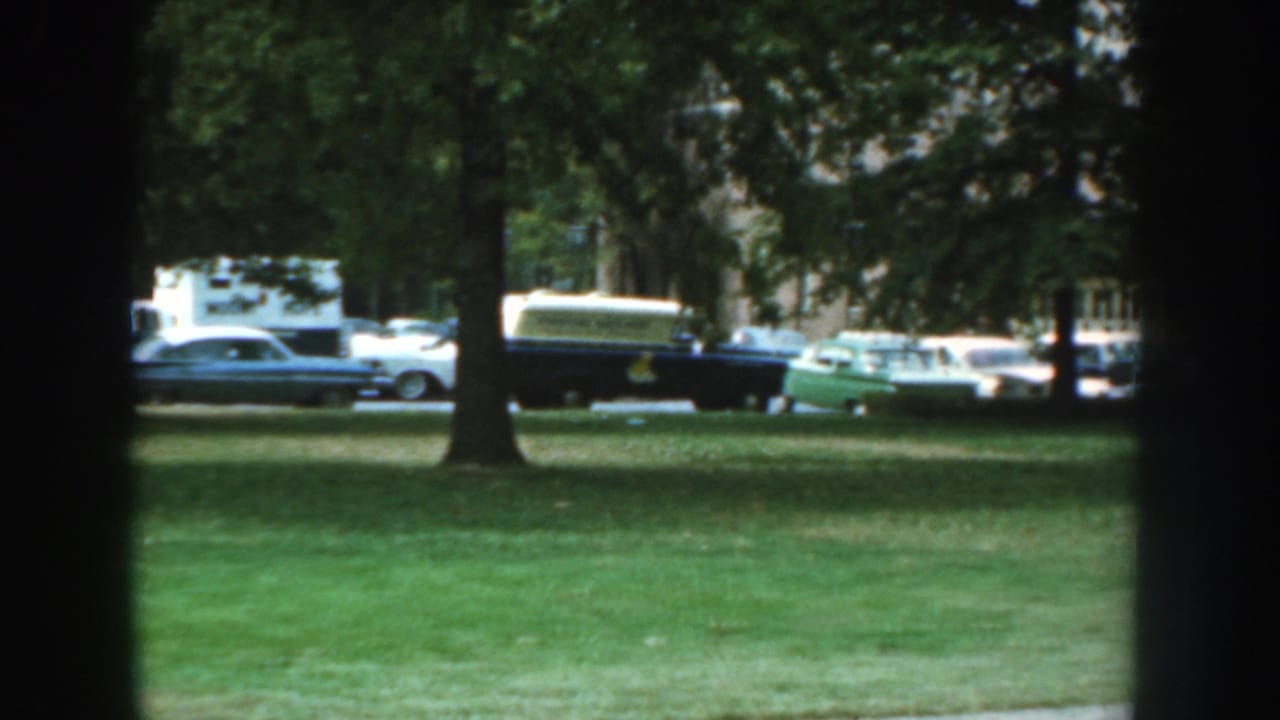 1959 Nebraska home-movie frame showing a lawn, trees, and parked cars near a brick residential building.