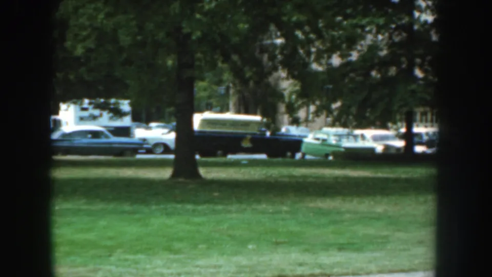 1959 Nebraska home-movie frame showing a lawn, trees, and parked cars near a brick residential building.