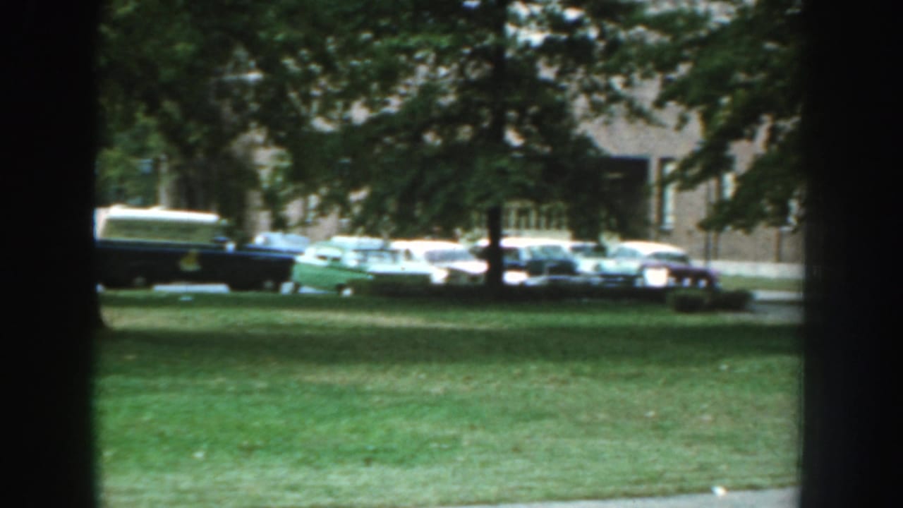 1959 Nebraska residential exterior with mature trees, grass, parked mid-century cars, and a brick apartment building.