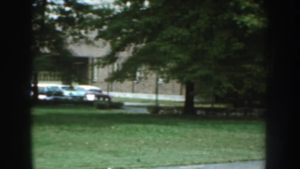 Home-movie view of a Nebraska apartment lawn with parked cars partly obscured by trees and dark foreground edges.