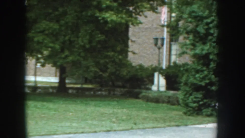 1959 Nebraska apartment-complex exterior with lawn, trees, shrubs, brick facade, and a visible American flag.