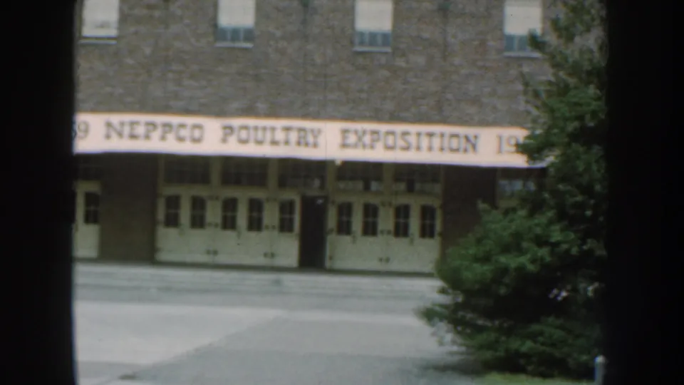 1959 Nebraska 8mm frame showing a brick building entrance with a NEPPCO Poultry Exposition banner partially framed by dark vertical foreground shapes.
