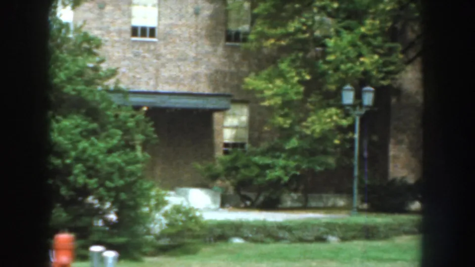 1959 Nebraska home-movie frame of the brick building grounds, showing trees, shrubs, lawn, and dark vertical foreground shapes during the pan.