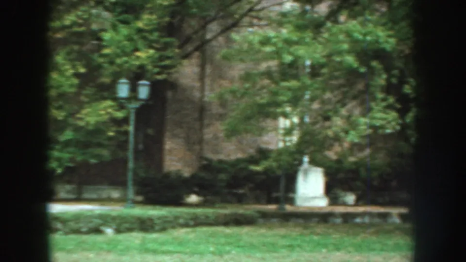 1959 Nebraska exterior frame showing streetlamps, trees, lawn, and the brick building facade as the handheld pan continues.