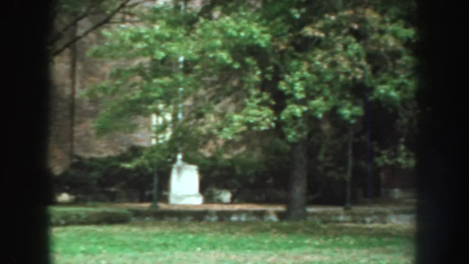 1959 Nebraska home-movie view of a grassy park-like area with mature trees, a white monument or marker, and dark foreground trunks.
