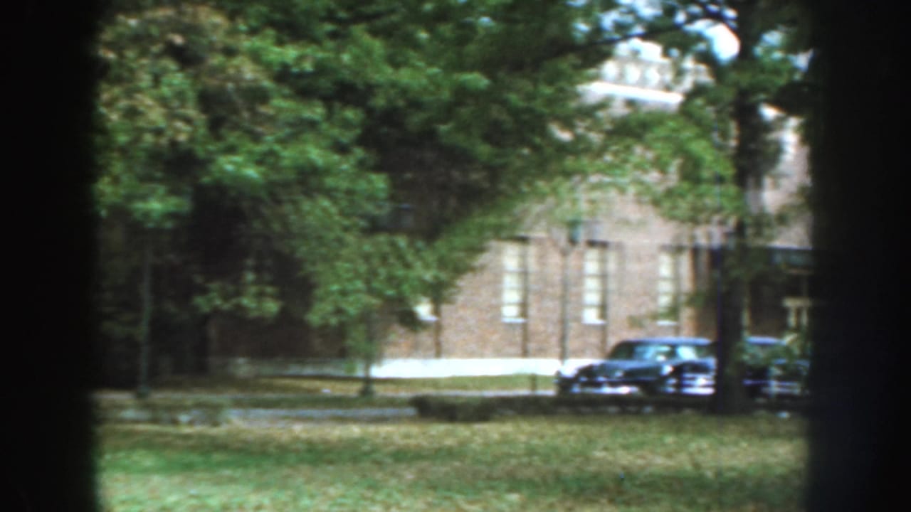 Trees frame a brick building and several late-1950s cars parked beside a lawn in Nebraska archival footage.