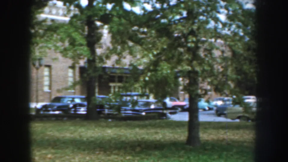 A row of classic American cars sits near a brick building, partly screened by trees in the 1959 Nebraska scene.