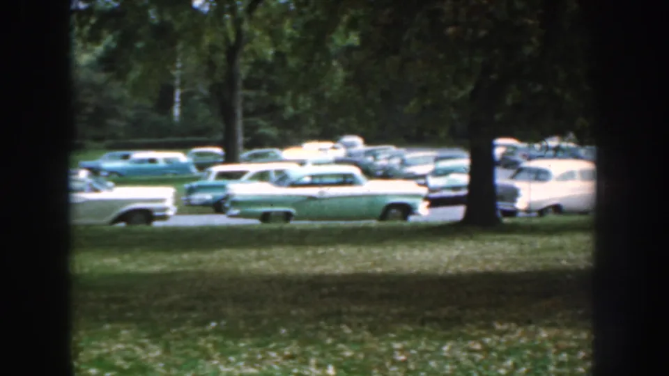 1959 Nebraska frame with multiple mid-century sedans and wagons parked along a road, partly obscured by trees.
