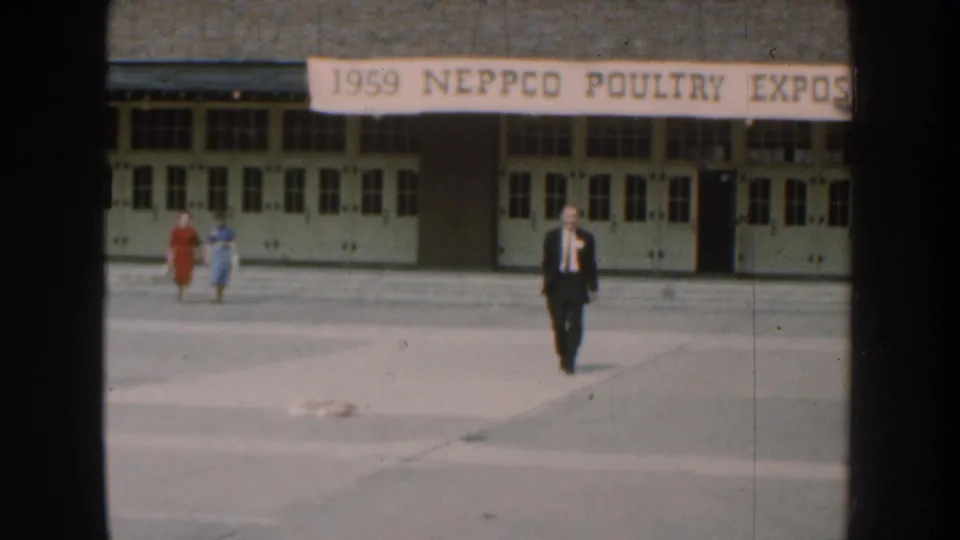 1959 Nebraska home-movie frame with a suited man approaching and two women walking near the poultry expo entrance.