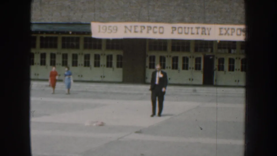 Nebraska 1959 8mm frame showing two women in colorful dresses and a suited man outside the poultry exposition building.