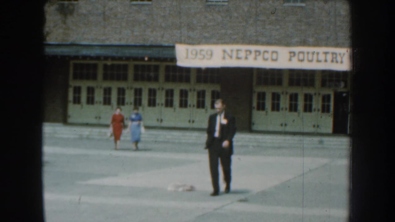 A man in a dark suit faces the camera as two women in dresses walk behind him outside the 1959 NEPPCO Poultry venue.