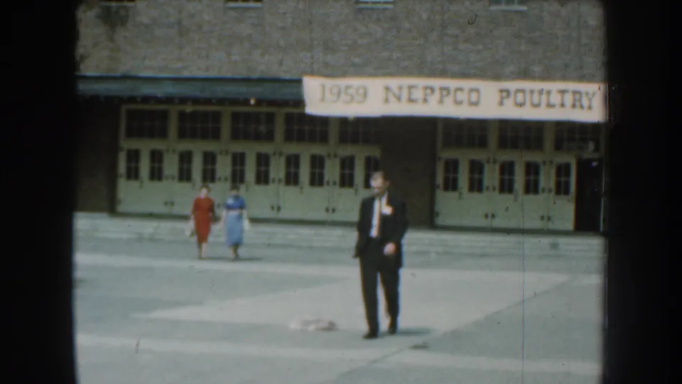 A man in a dark suit faces the camera as two women in dresses walk behind him outside the 1959 NEPPCO Poultry venue.
