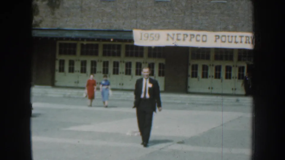 The suited man moves closer across the paved area, with two women in colorful dresses behind him at the Nebraska poultry-event setting.