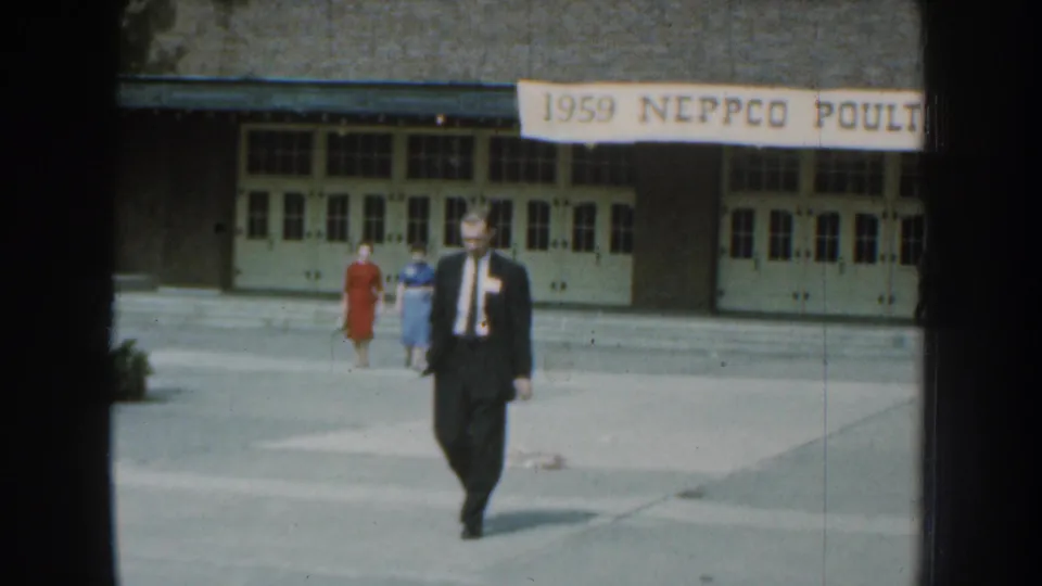Near the end of the clip, the suited man fills more of the frame as two women walk behind him at the 1959 NEPPCO Poultry site.