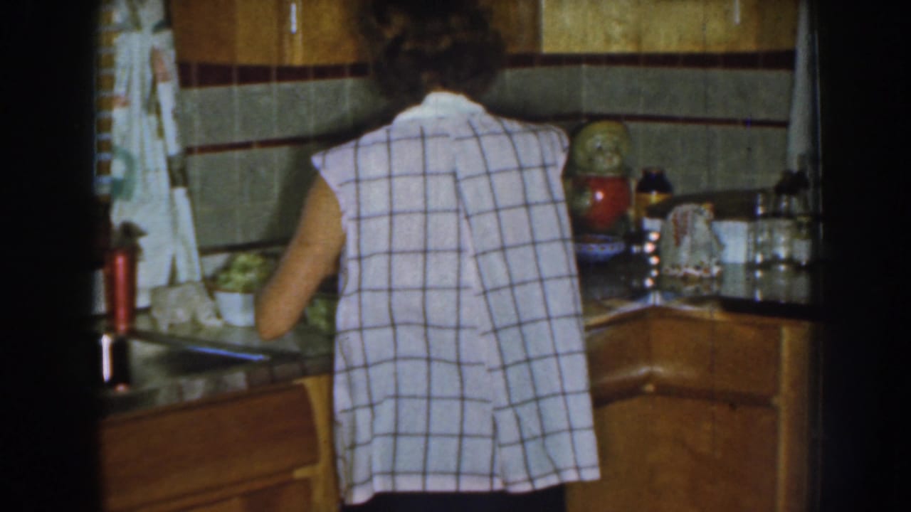 Woman seen from behind working at a kitchen counter beside wood cabinets and a tiled backsplash.