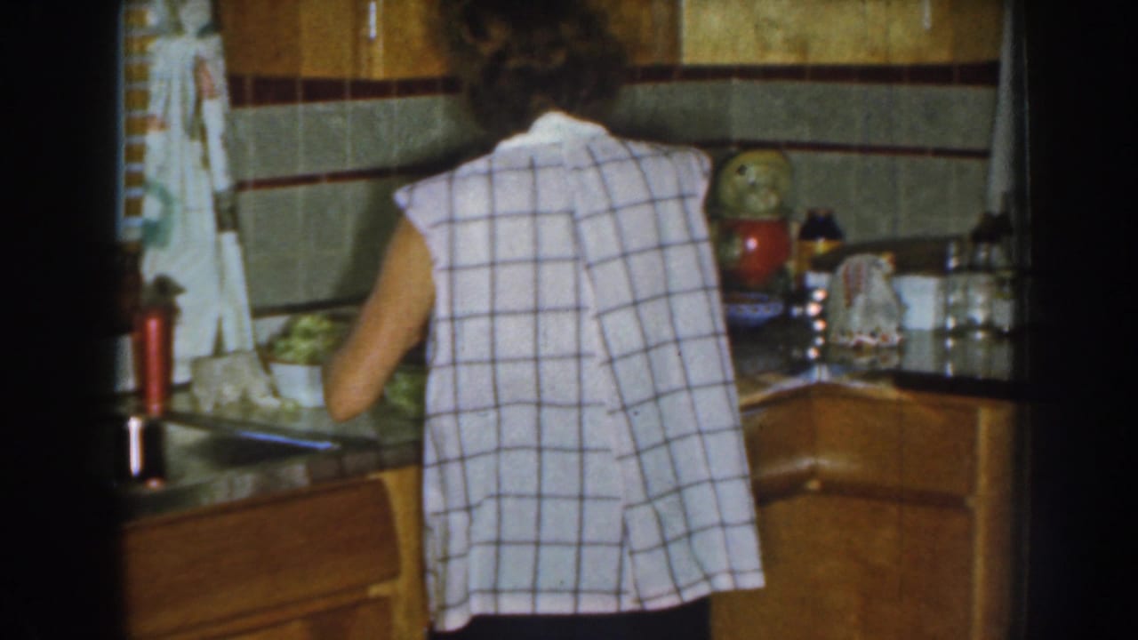 A woman prepares food at a counter in a 1959 Nebraska kitchen, with wood cabinetry, tiled walls, curtains, and small containers visible.