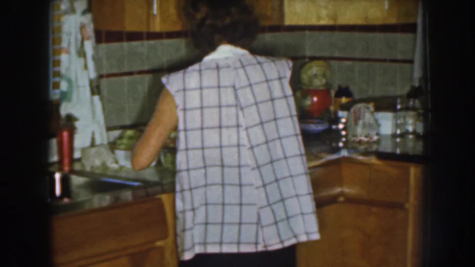 The 1959 Nebraska home-movie frame shows a woman from behind at a kitchen counter, surrounded by wood drawers, tile, curtains, and utensils or containers.
