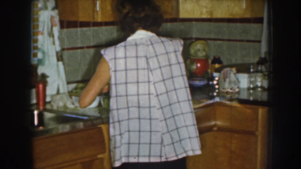 A woman in a sleeveless plaid shirt remains at the counter in a 1959 Nebraska kitchen, with wood cabinets, tiled backsplash, curtains, and household items visible.