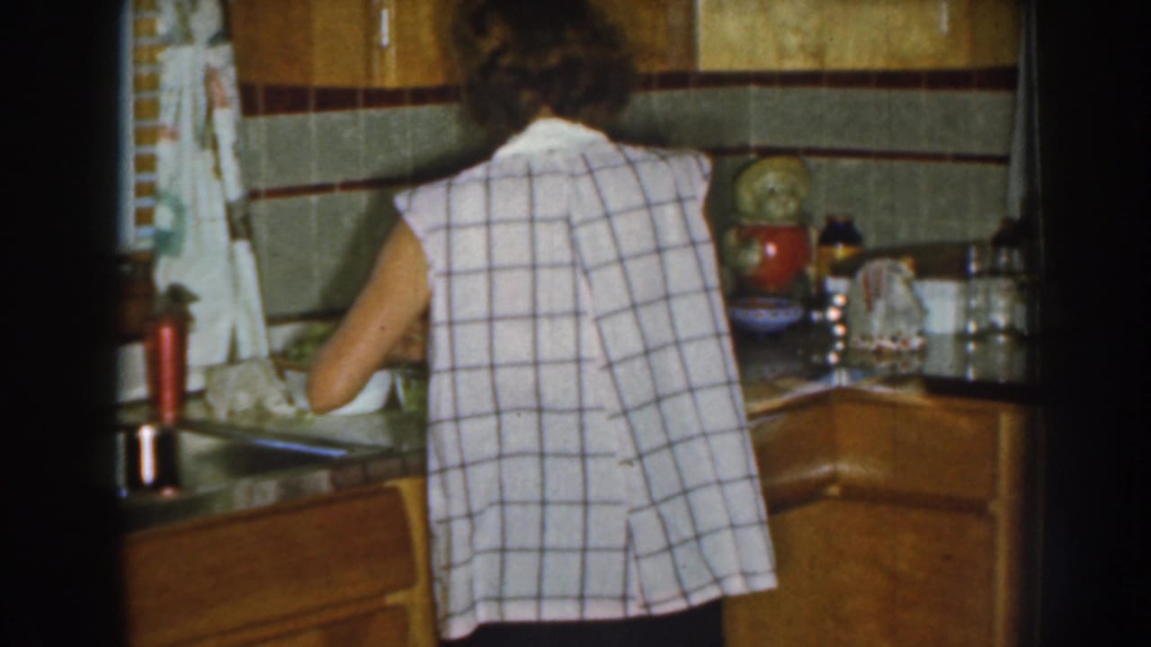 Woman seen from behind working at a kitchen counter beside wood cabinets and tiled walls.