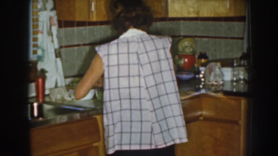 In a 1959 Nebraska home kitchen, a woman in a checked sleeveless dress works at the counter with wood cabinets and tiled walls nearby.
