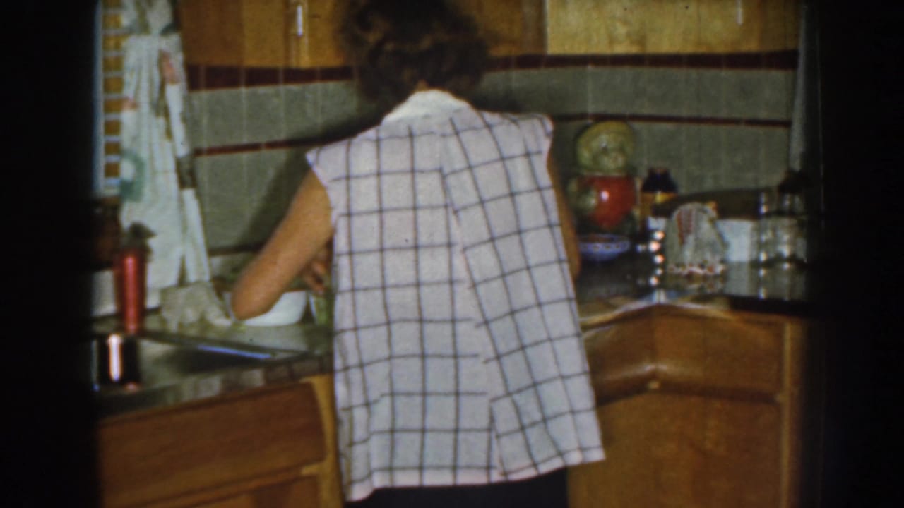 Woman at the counter in a kitchen with cabinets, tile, containers, and a red thermos.