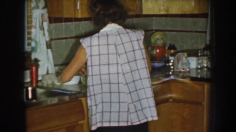 The woman continues working at the counter in the Nebraska kitchen, with tiled walls, wood cabinetry, and a red thermos visible.
