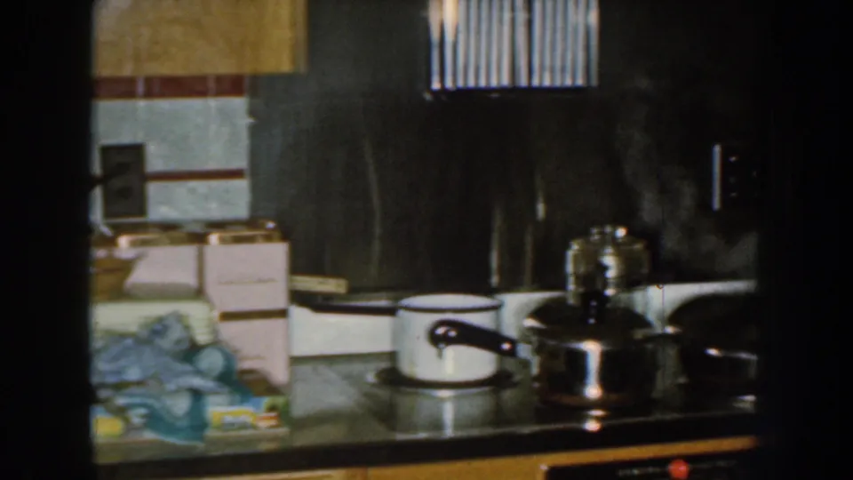 The camera frames the stove area of the 1959 Nebraska kitchen, with pots, containers, tiled wall, and dark appliances visible.