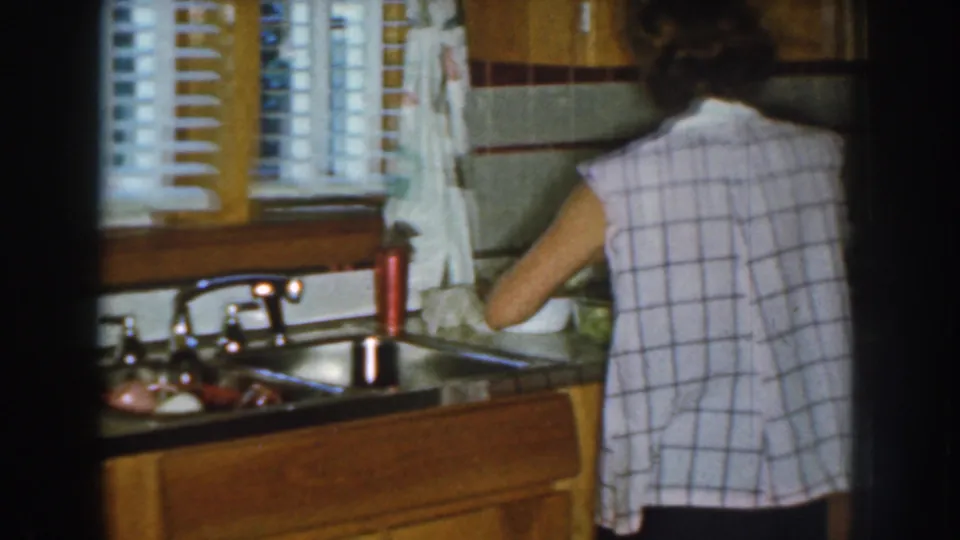 A woman in a sleeveless checked shirt works at the kitchen sink in 1959 Nebraska home-movie footage.