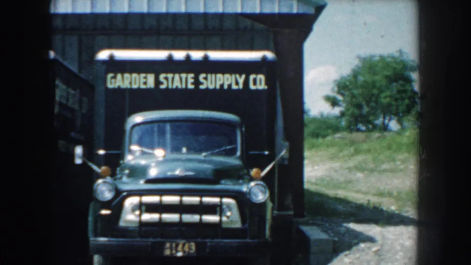 1959 Nebraska home-movie frame with a dark green Garden State Supply Co. truck facing the camera beside metal-sided structure.