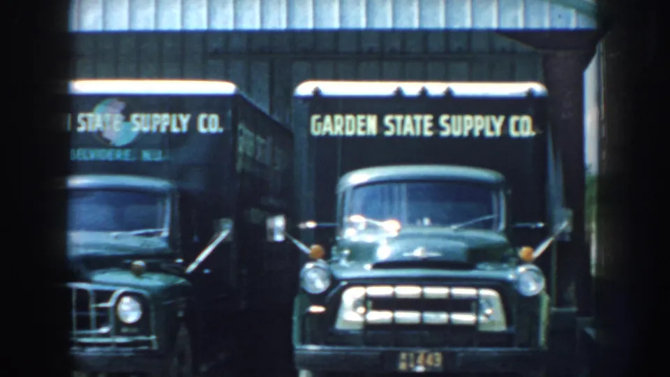 A 1959 Nebraska frame shows Garden State Supply Co. trucks parked beside a shed-like building, with bright 8mm exposure and soft focus.