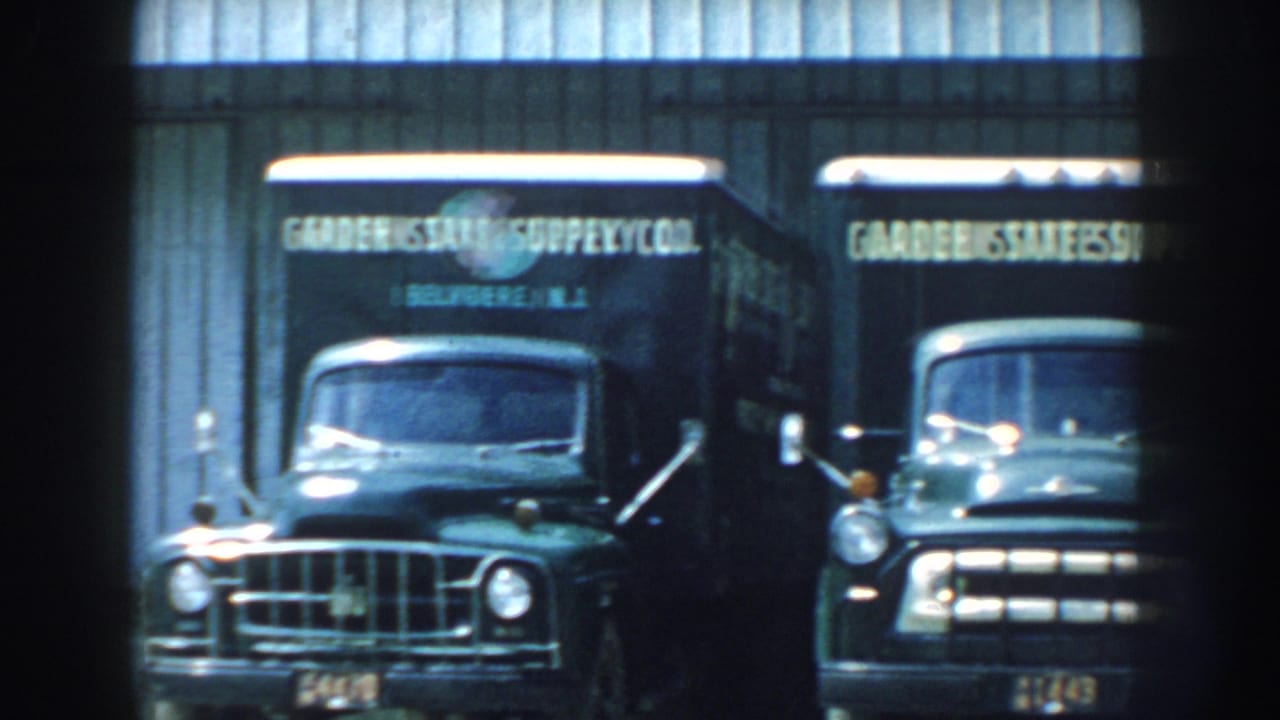 Two dark green delivery trucks parked outside a pale corrugated metal building.