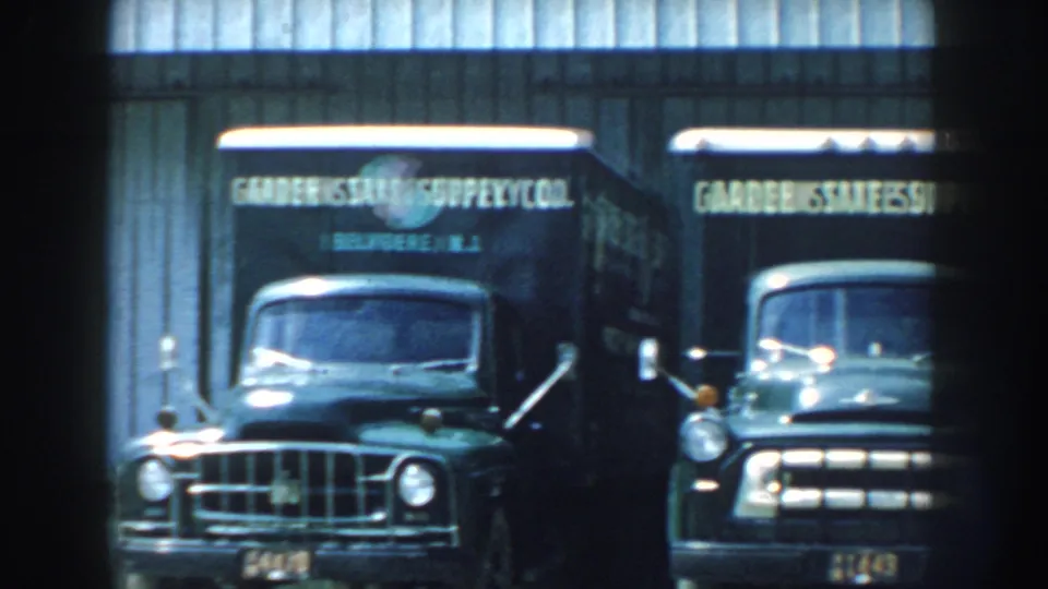 1959 Nebraska 8mm frame showing two Garden State Supply Co trucks beside a light corrugated building.