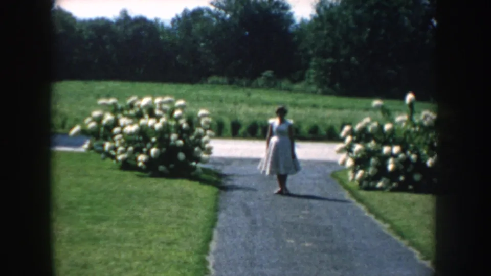 In 1959 Nebraska home-movie footage, a woman in a light dress walks along a paved driveway bordered by lawns and white flowering shrubs.