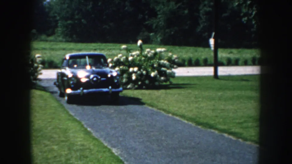 The black mid-century car moves closer along the driveway in 1959 Nebraska, framed by bright lawn, shrubs, and dark film edges.
