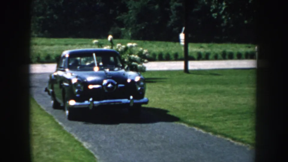A black vintage car reaches the foreground of a Nebraska driveway, with lawns, white flowering shrubs, and trees visible behind it.