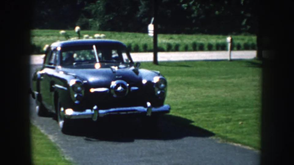 1959 Nebraska 8mm frame showing a dark blue mid-century sedan entering a residential driveway from near a window view.
