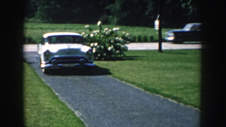 1959 Nebraska 8mm frame showing a white mid-century car approaching along the driveway beside flowering shrubs.