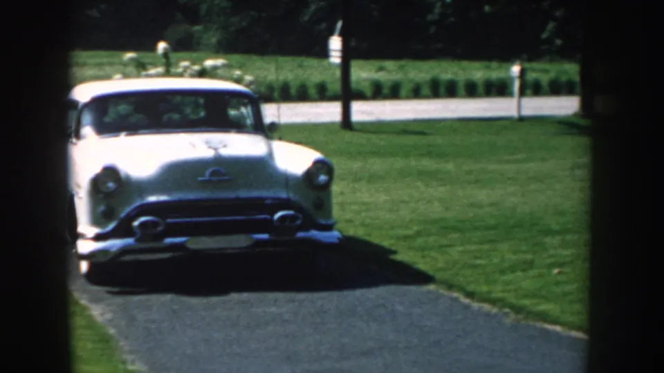 1959 Nebraska home-movie frame with a white mid-century car stopped or approaching near the camera on a residential driveway.