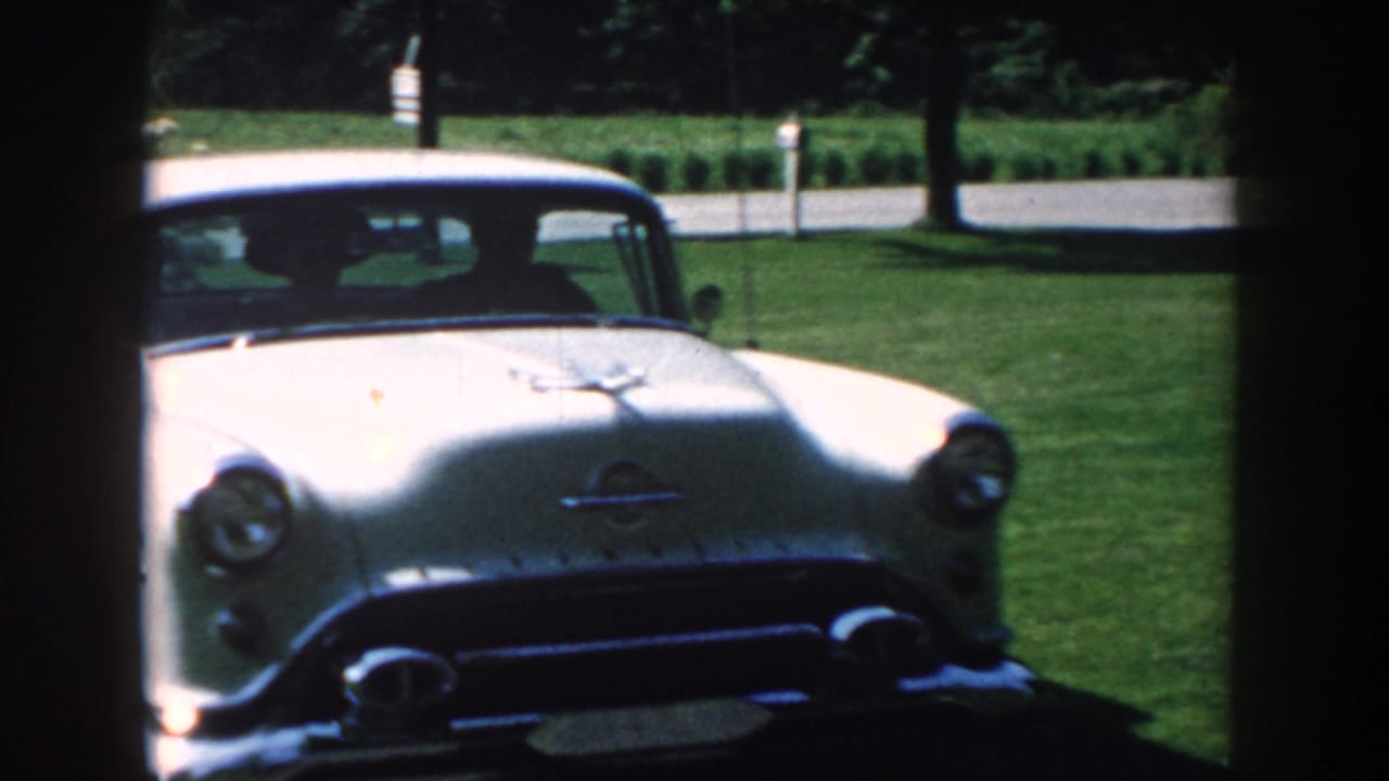 White 1950s car parked beside a lawn and garden shrubs.