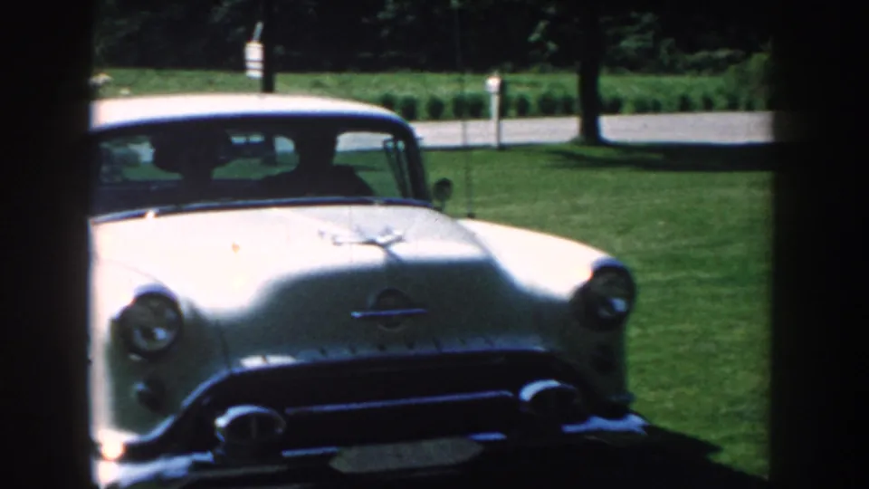 White mid-century car near a green lawn and garden area in 1959 Nebraska home-movie footage.