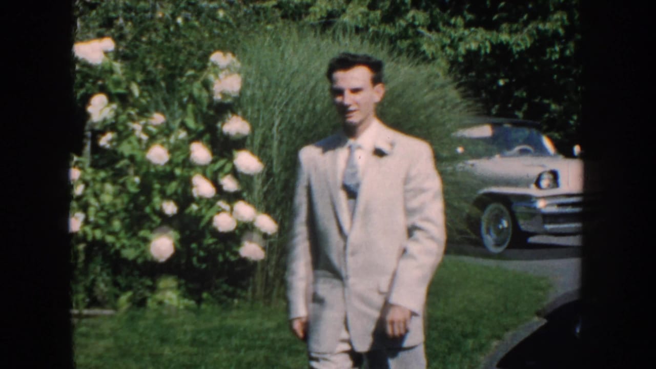 Man in a light suit walks across a garden lawn near flowers and a parked car.