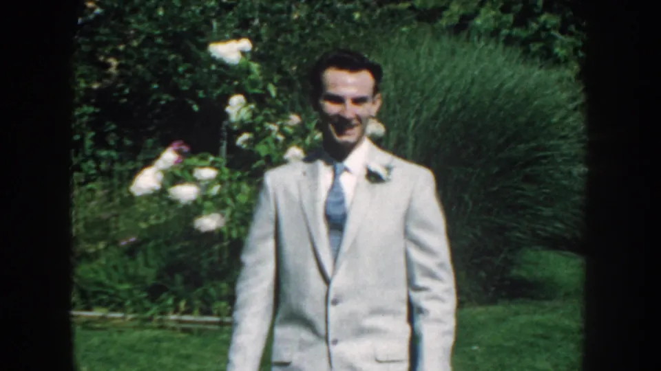 Smiling suited man with a boutonniere stands in a lush Nebraska garden in 1959 home-movie footage.