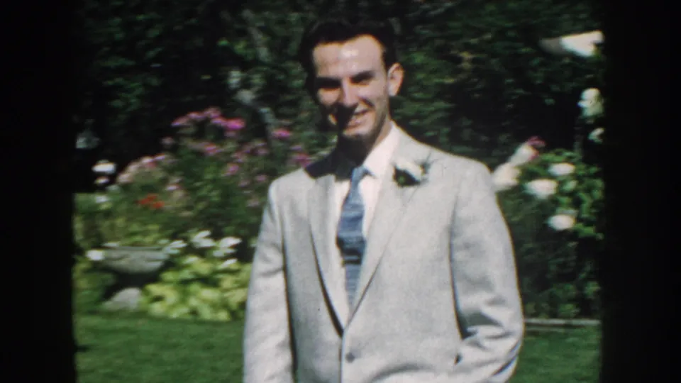 Close view of a smiling man in a pale suit and tie in a 1959 Nebraska garden scene.