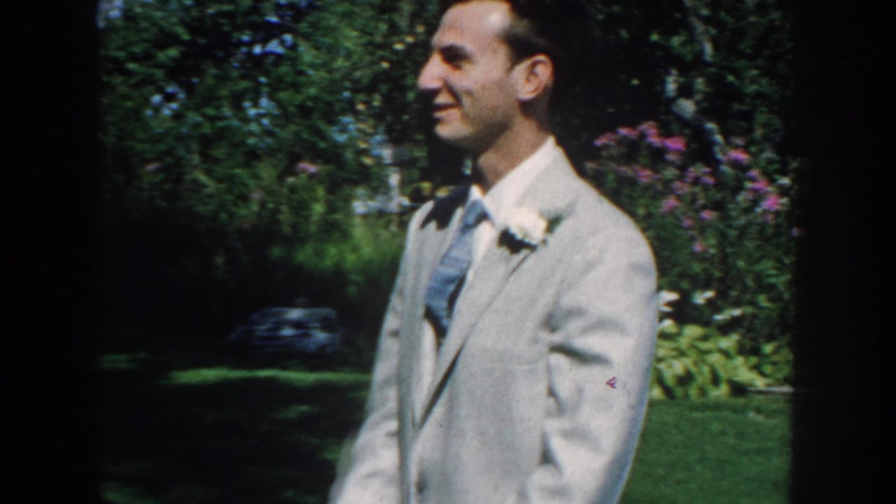 Smiling man in a light grey suit standing outdoors near a garden.