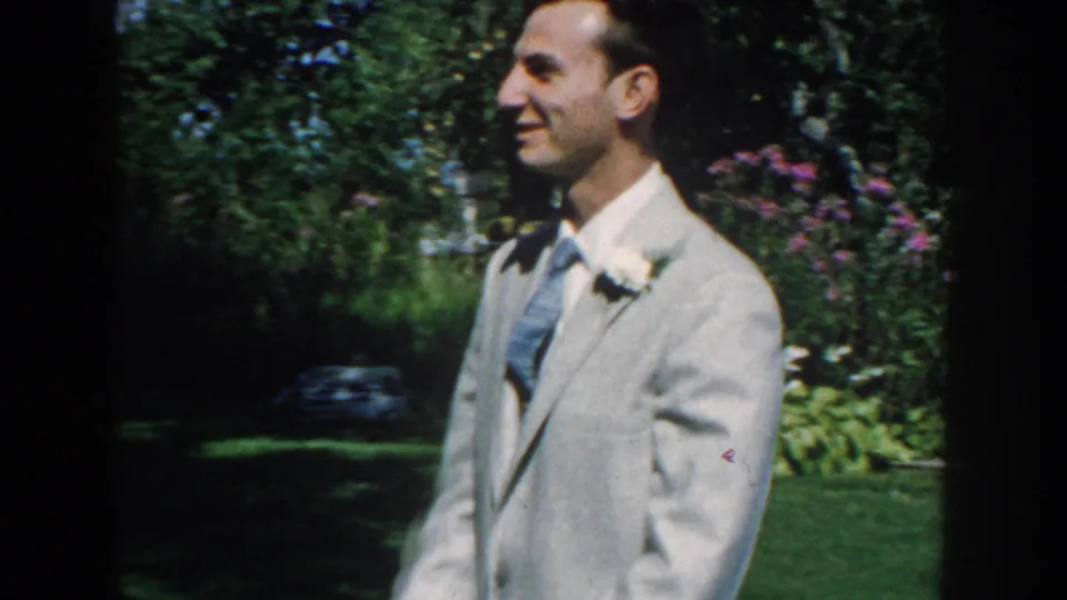 In Nebraska 1959 home-movie footage, a smiling man in a light grey suit stands near flowering shrubs and lawn.