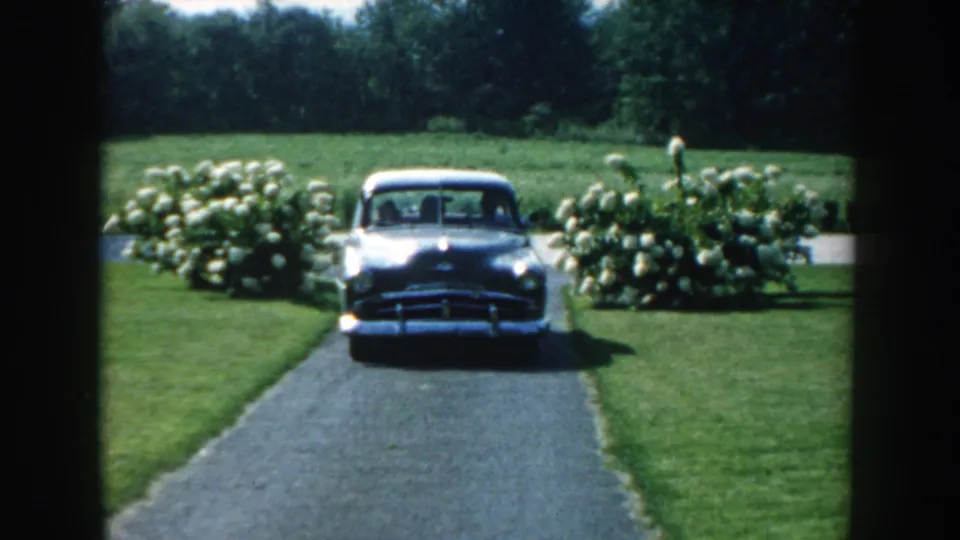1959 Nebraska home-movie frame of a gray mid-century sedan driving toward the camera on a landscaped driveway.