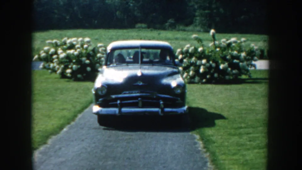 The gray car nears the camera on a Nebraska garden driveway, with chrome trim catching sunlight in the 8mm frame.