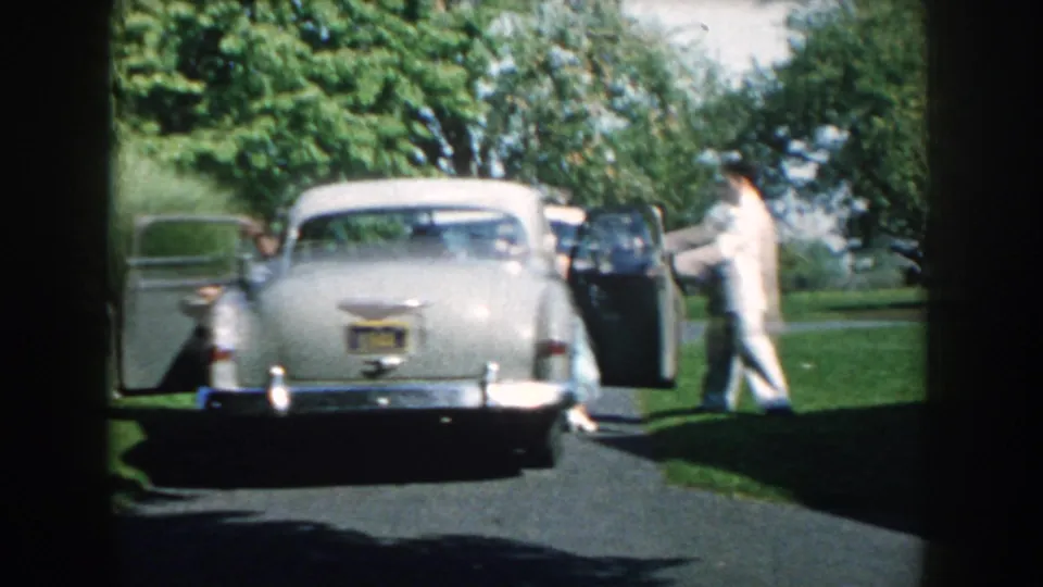 The sedan has stopped on the driveway, with doors open and a suited man standing near the passenger side in the garden setting.